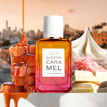 Queens Caramel 50ml Eau De Parfum on white table with block of caramel with melted caramel being poured on top, whipped cream, pink salt, and pieces of white chocolate, with sunset city scene in background.
