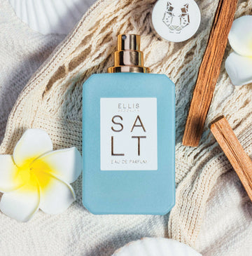 SALT Eau de Parfum 50ml sans bouchon posé sur une couverture blanche en tissu tissé à côté de fleurs blanches et jaunes, bouchon avec un motif léopard argenté, et des bâtons de bois