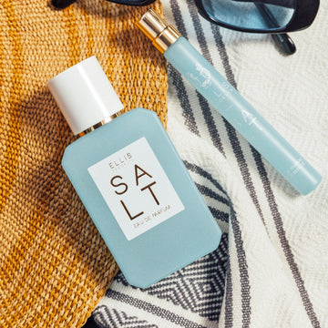 SALT Eau de Parfum 50ml et 10ml posés sur des serviettes blanches, grises et jaunes à côté de lunettes de soleil noires avec des verres bleus