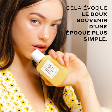 Femme aux cheveux bruns portant un haut blanc avec des fleurs jaunes tient SWEET Eau de Parfum 50ml contre son menton sur un fond blanc cassé avec du texte noir qui dit « cela evoque le doux souvenir d'une epoque plus simple. »