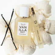 Gousses de vanille, sel de vanille 50 ml Eau de Parfum et sel de vanille 10 ml Eau de Parfum en vaporisateur de voyage reposant sur des boules de glace à la vanille, sur fond blanc.