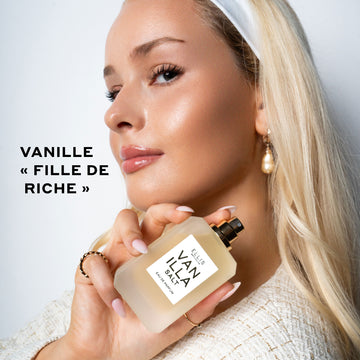 Mannequin aux cheveux blonds et vernis à ongles nude portant un serre-tête blanc, une boucle d'oreille en perle, des bagues en or et une chemise en bouclé crème tenant le parfum Vanilla Salt 50ml sans son bouchon près de l'épaule sur fond blanc avec un texte noir indiquant 
