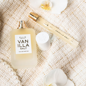Vanilla Salt 50ml Eau de Parfum avec le bouchon retiré à côté du Vaporisateur de Voyage Vanilla Salt 10ml Eau de Parfum, fleurs blanches et collier de perles, sur un tissu bouclé crème.