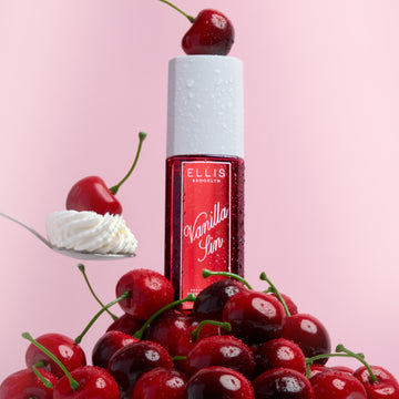 Brume parfumée VANILLA SIN 240ml avec une cerise posée sur le bouchon, posée sur un tas de cerises et une cuillère avec une cerise sur un tas de crème fouettée sur un fond rose