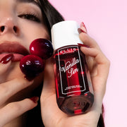 Un modèle aux cheveux bruns et aux ongles rouges regarde la caméra tout en tenant le parfum en brume VANILLA SIN 90ml et deux cerises contre sa joue sur un fond rose