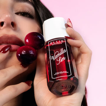 Un modèle aux cheveux bruns et aux ongles rouges regarde la caméra tout en tenant le parfum en brume VANILLA SIN 90ml et deux cerises contre sa joue sur un fond rose