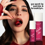 Un mannequin aux cheveux bruns et aux ongles rouges portant un t-shirt rouge et des boucles d'oreilles en or mange une cerise sur un fond blanc avec des bouteilles de VANILLA SIN 240ml et 90ml et un texte noir et blanc qui dit « ca sent la soiree à Brooklyn. »