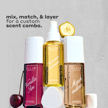 VANILLA SIN Perfume Mist 240ml next to a cherry, BANANA Milkshake Perfume Mist 240ml sitting on top of it's cap, MARSHMALLOWS Perfume Mist 240ml with a slice on banana on top of it and a marshmallow next to it, all covered in water droplets on a gray, rough background with black text that reads "mix, match, & layer for a custom scent combo."