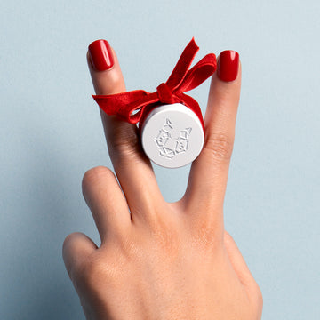 Model with red nails holding 50ml Eau De Parfum cap wrapped in red velvet ribbon between two fingers against blue background.