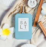 SALT Eau de Parfum 50ml without cap lays on white woven blanket next to white and yellow flowers, cap with silver leopard design, and sticks of wood