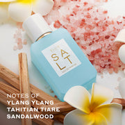 SALT Eau de Parfum 50ml lays next to white and yellow flowers, pink salt, and sticks of wood against white background with white text that reads "Notes of ylang ylang, Tahitian tiare, sandalwood'