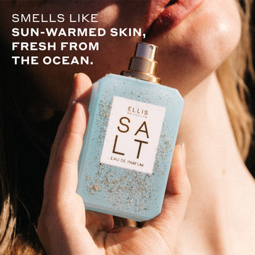 Woman with blonde hair holds SALT Eau de Parfum 50ml without cap covered in sand against her chin bathed in sunlight with white text that reads "Smells like sun-warmed skin, fresh from the ocean."
