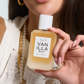 Woman with brown hair wearing gold hoop earrings, gold and pearl necklaces, and a white embroidered tank top holds VANILLA SALT Eau de Parfum 50ml under her chin.
