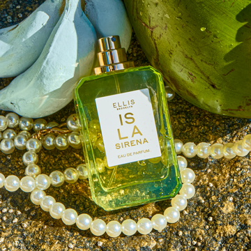 ISLA SIRENA Eau de Parfum 50ml without cap lays on white pearls and a bunch of blue bananas next to a green coconut on a rocky surface