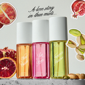 NEW CRUSH Perfume Mist 90ml, LOVE POM Perfume Mist 90ml, and FOREVER PISTACHIO Perfume Mist 90ml stand on white marble between sliced pomegranate, sliced blood orange, and pistachios against a gray background with drawn images of sliced pomegranate, sliced blood orange, and pistachios with a white outline with black text that reads "A love story in three mists..."