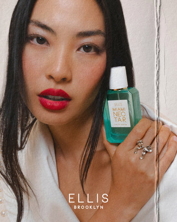 Model with brown hair wearing red lipstick, white shirt, and silver, bow ring holding Miami Nectar Eau de Parfum 50ml against her cheek on white background with fold on right side and white text that reads "Ellis Brooklyn"