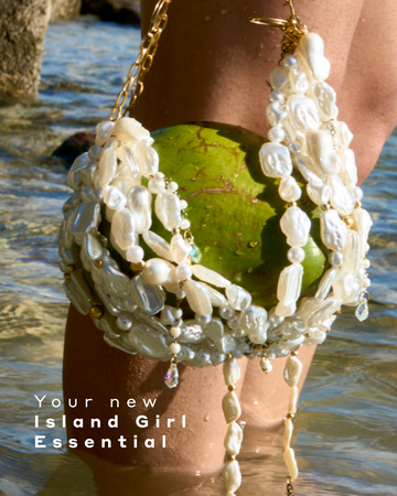 Model standing in ankle deep clear ocean water holds a green coconut in a white, shell bag with gold hardware next to their knees with white text that reads "Your new Island Girl Essential"