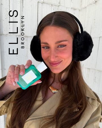 Woman with brown hair wearing black fuzzy earmuffs, a tan trench coat, and a gold necklace holds MIAMI NECTAR Eau de Parfum 50ml with black text that reads "Ellis Brooklyn" in front of a white wall