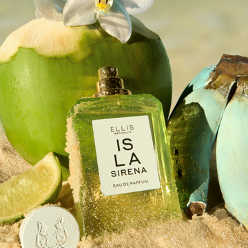 ISLA SIRENA Eau de Parfum 50ml without cap with sand on the bottle sits in sand next to it's cap, a slice of lime, a bunch of blue bananas, and a green coconut with a white flower on it