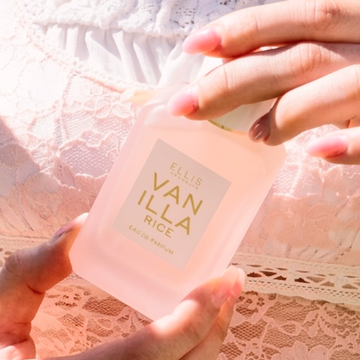Closeup of model with pink manicure holding VANILLA RICE Eau de Parfum 50ml against white, lace shirt