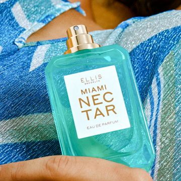 Closeup of model holding MIAMI NECTAR Eau de Parfum 50ml without cap against a blue, striped, sparkly dress