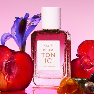 PLUM TONIC Eau de Parfum 50ml next to sliced plums, amber stones and a purple iris covered in water droplets on a pink background 