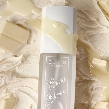 Closeup of COCOA BLANCO perfume mist 90ml laying on vanilla icing, chunks of white chocolate, and a drizzle of white chocolate across the bottle. 