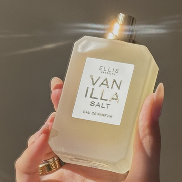 Hand with nude manicure and gold rings holds VANILLA SALT Eau de Parfum 50ml without cap against a gray background with light reflection