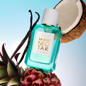 Miami Nectar 50ml Eau De Parfum resting on pink pinapple, vanilla beans, and half coconut, on blue background.