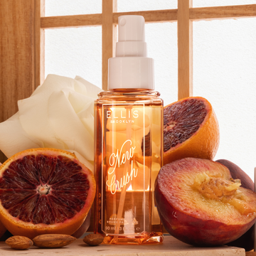 NEW CRUSH Perfume Mist 90ml without cap next to blood orange halves, nectarine halves, almonds, and a white flower on a tan surface against a window with six panes and tan, wooden muntins 