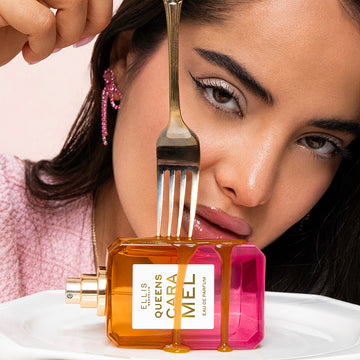 Model with black hair wearing pink diamond earring, gold necklace, and pink shirt holding fork to Queens Caramel 50ml Eau De Parfum bottle without cap, covered in melted caramel on white plate, on white table, on light pink background.