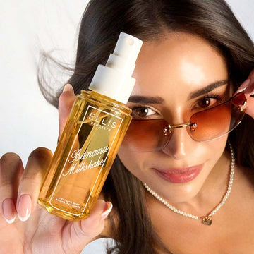 Model with brown hair and a French manicure wearing brown sunglasses and a pearl necklace looks up at the camera while holding BANANA MILKSHAKE 90ml perfume mist without a cap on a white background
