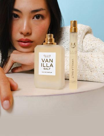 Model with brown hair wearing white and gold jacket resting face on hands  on top of white table behind Vanilla Salt 50ml Eau De Parfum without cap and Vanilla Salt 10ml Eau De Parfum Travel Spray, on blue background.