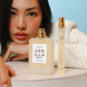 Model with brown hair wearing white and gold jacket resting face on hands  on top of white table behind Vanilla Salt 50ml Eau De Parfum without cap and Vanilla Salt 10ml Eau De Parfum Travel Spray, on blue background.