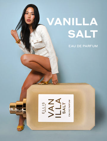 Model with black hair wearing white jacket and shorts, gold sandals, and gold bracelets resting one foot on oversized bottle of Vanilla Salt Eau De Parfum 50ml without cap, and other foot on ground, on blue background with white text that reads "Vanilla Salt Eau De Parfum".