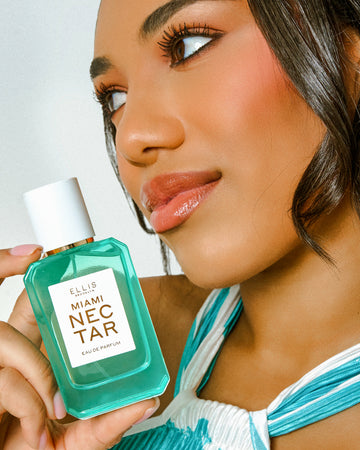 Woman with brown hair wearing blue and white halter deck dress holds MIAMI NECTAR Eau de Parfum 50ml on a white background