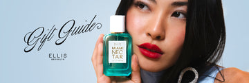 Model with brown hair wearing red lipstick, blue dress, and diamond clip holding Miami Nectar 50ml Eau De Parfum to face, on blue background with black text that reads "Gift Guide" above black Ellis Brooklyn logo.