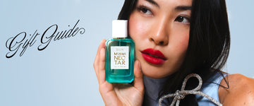 Model with brown hair wearing red lipstick, blue dress, and diamond clip holding Miami Nectar 50ml Eau De Parfum to face, on blue background with black text that reads "Gift Guide".