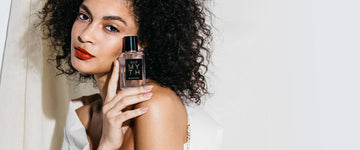 Model with black curly hair, nude nail polish, red lipstick, wrapped in white sheet against white fabric background holding Myth 50ml Eau De Parfum on shoulder.