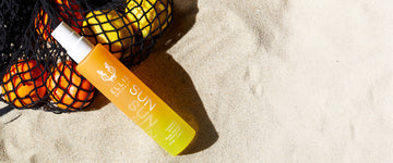 Sun Hair and Body Fragrance Mist 100ml in sand leaning on black mesh bag filled with oranges and lemons.