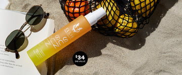 Sun Hair and Body Fragrance Mist 100ml in sand leaning on black mesh bag filled with oranges and lemons, and corner of open book, next to sunglasses, and black circle with white text that reads