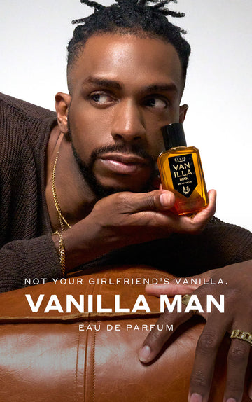 Model with brown hair weaing gold necklaces, bracelets, rings, and brown sweater holding Vanilla Man 50ml Eau De Parfum in one hand next to face while leaning on brown leather couch, on white background above white text that reads "Not Your Girlfriend's Vanilla. Vanilla Man Eau De Parfum"