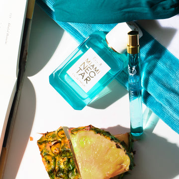 Miami Nectar 50ml Eau De Parfum and Miami Nectar 10ml Eau De Parfum Travel Spray leaning on blue and white striped folded towel next to bikini top, pineapple, and open book, on white background.