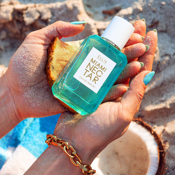 Model with teal nail polish and sand on hands wearing gold bracelet hold Miami Nectar 50ml Eau De Parfum and pineapple in both hands against sand background with blue and white striped towel, and coconut.