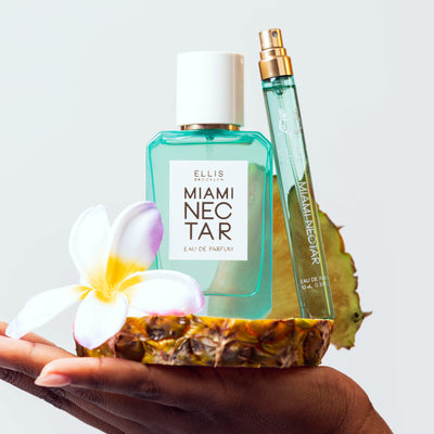 Model with nude nail polish holding Miami Nectar 50ml Eau De Parfum, Miami Nectar 10ml Eau De Parfum Travel Spray without cap, and pink, white, and yellow flower on slice of pineapple on white background