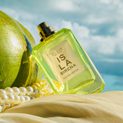 ISLA SIRENA Eau de Parfum 50ml without cap leans against a green coconut and white pearls on top of tan fabric against a blue sky and white cloud background