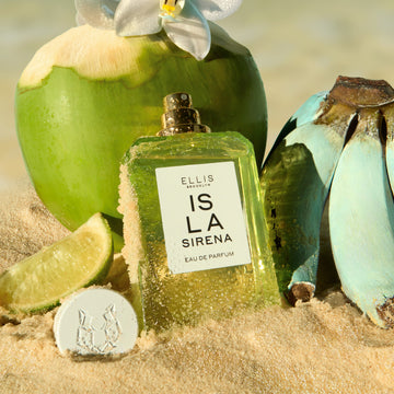 ISLA SIRENA Eau de Parfum 50ml sans bouchon avec du sable sur le flacon, posé dans le sable à côté de son bouchon, une tranche de citron vert, une grappe de bananes bleues et une noix de coco verte avec une fleur blanche dessus