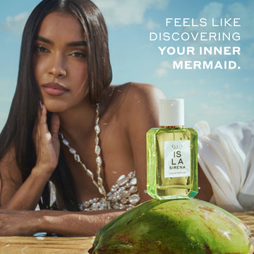 Model with brown hair wearing a white shell top and white skirt lays on her stomach in wet sand with her hand on her chin behind ISLA SIRENA Eau de Parfum 50ml sitting on top of a green coconut with white text that reads "Feels like discovering your inner mermaid." all with a blue sky and white cloud background.