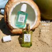 One 50mL bottle of ISLA SIRENA eau de parfum placed on sand with the white cap off bottle and set to the left and one 50mL bottle of MIAMI NECTAR eau de parfum placed in a halved coconut. On the right there is text that reads 