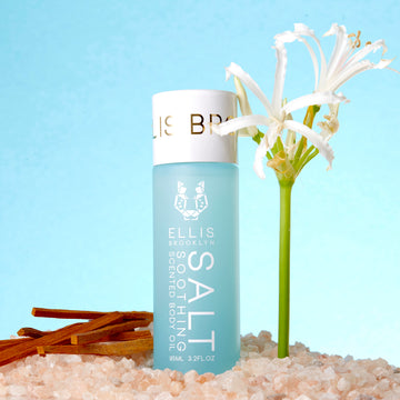Huile corporelle parfumée apaisante Salt 95 ml au sel rose à côté d'une fleur blanche et du bois de santal, sur fond bleu.